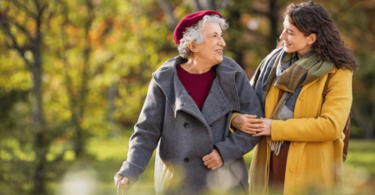 Elderly woman and young support worker laughing and walking together in a sunny outdoor park